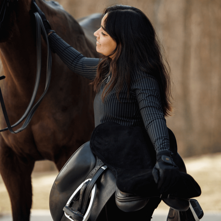 Woman wearing Bella pull on riding leggings turned toward her horse smiling while holding her dressage saddle. 