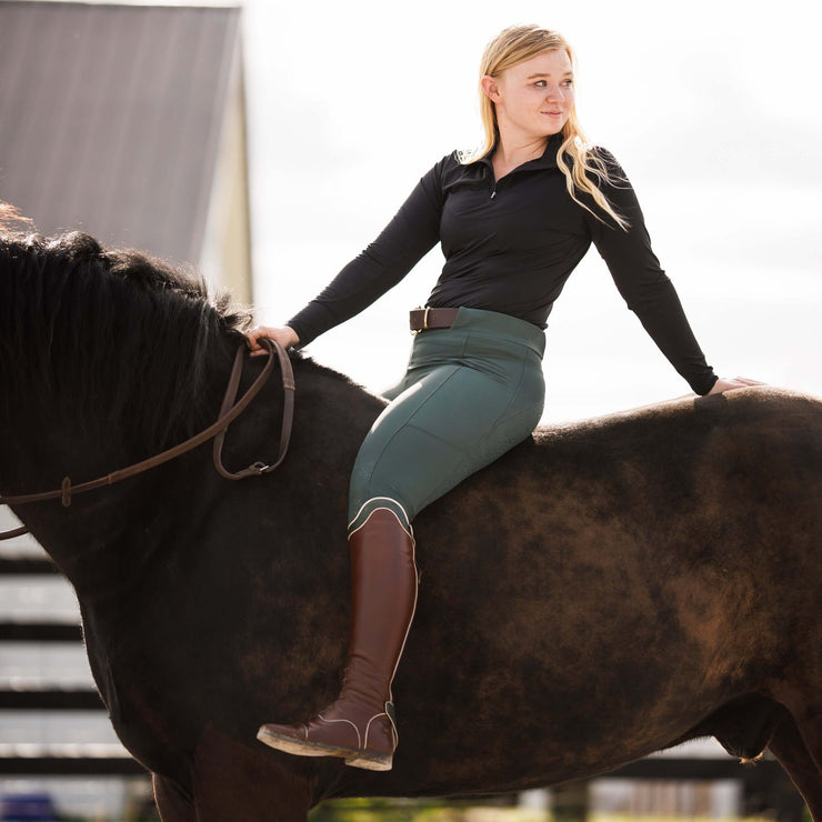 Woman on a bay horse wearing ribbed hybrid pull on breeches and brown tall boots. The waistband has two long belt loops.