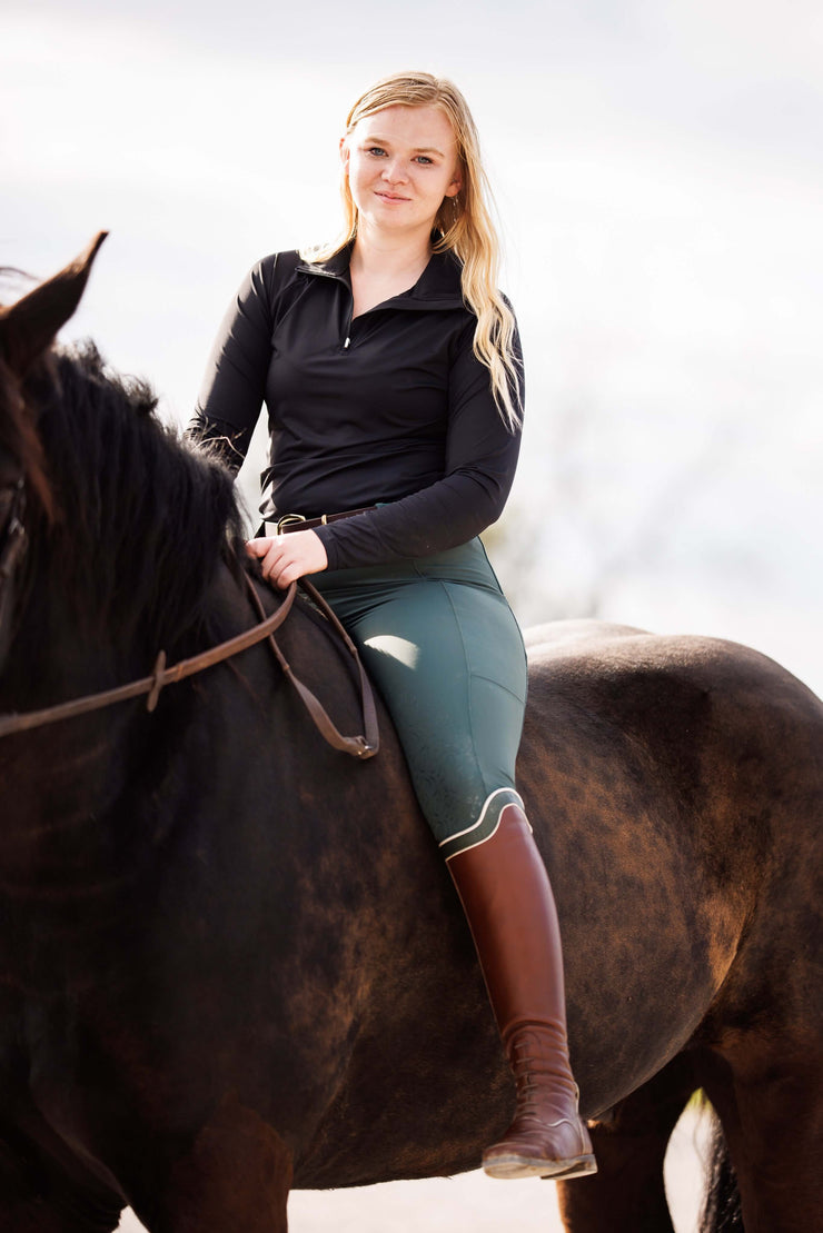 Woman on a bay horse wearing ribbed hybrid pull on breeches and brown tall boots. The waistband has two long belt loops.