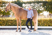 Woman inside equestrian arena wearing May Babes riding breeches in black. She has her hand on her horses withers and is looking at him smiling. She is wearing a blue button up blouse and sun hat. 