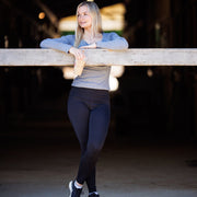 Woman wearing black Phoenix legggings with her legs crossed while standing and learning against a board inside a barn. She is wearing a grey long sleeve shirt that highlights the high waist of the leggings, and holding a coffee in her right hand while looking off into the distance. 