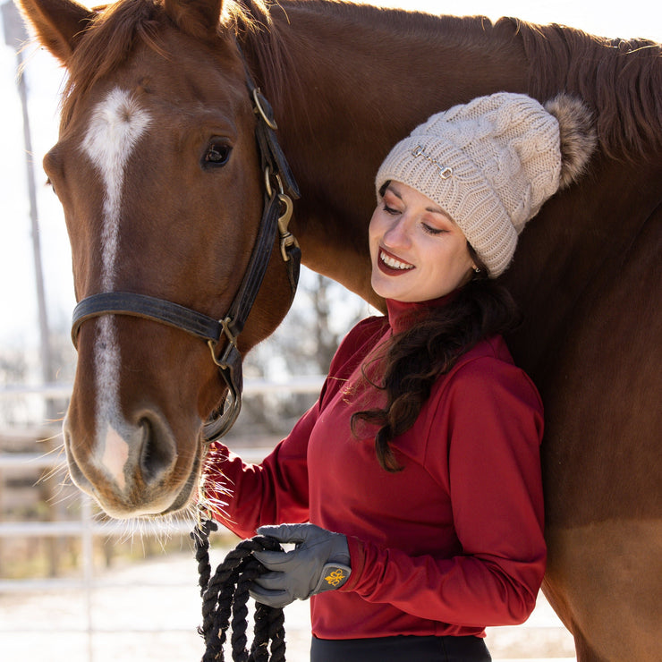 Woman in a red GG Technical shirt and black May Babes leggings stands with her horse in the snow. She holds a cup of coffee and is wearing a tan knitted beanie while smiling at her horse