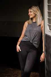 Woman in black riding legggings and grey tank top, leans agains the door of a horse trailer with her right hand in her right pocket. 